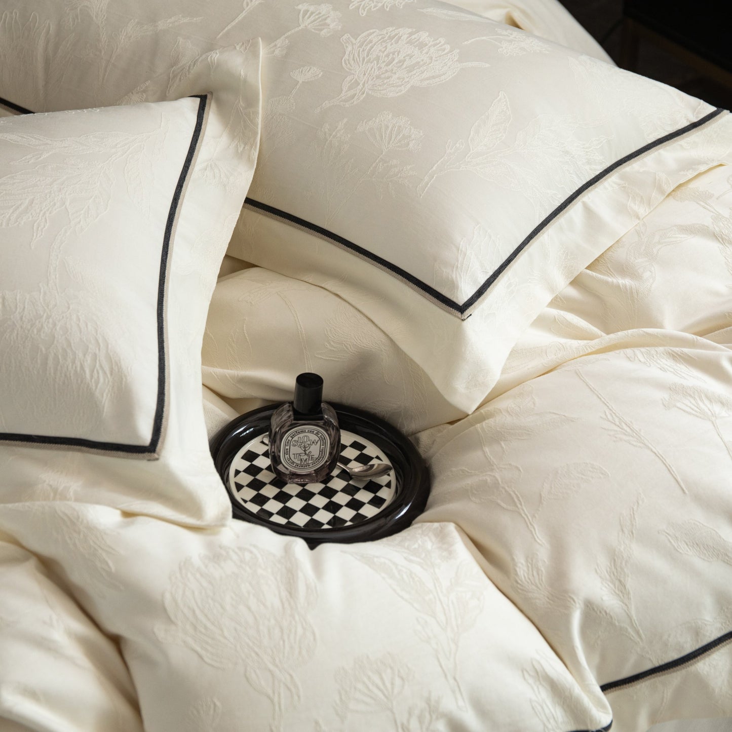 Cream sateen bedding set with subtle floral patterns and black trim, styled with a small decorative tray and bottle.