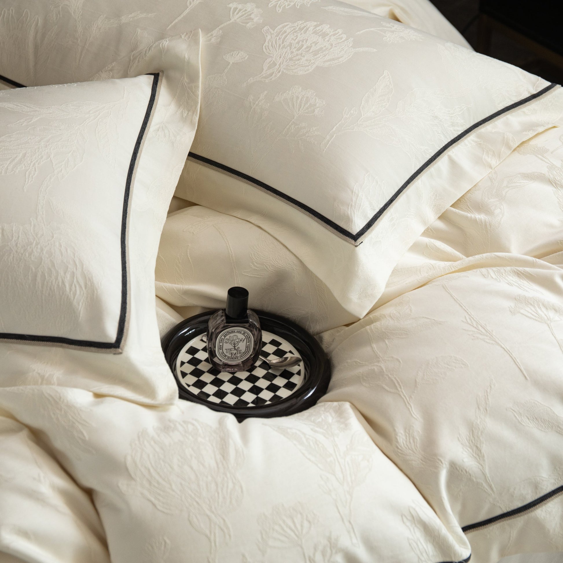 Cream sateen bedding set with subtle floral patterns and black trim, styled with a small decorative tray and bottle.